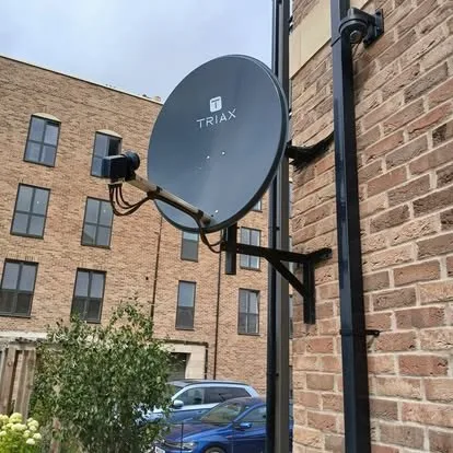A satellite dish on a bracket against brick, with an apartment block behind.