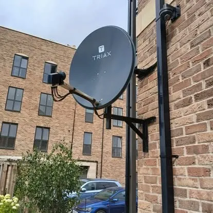 A satellite dish on a bracket against brick, with an apartment block behind.